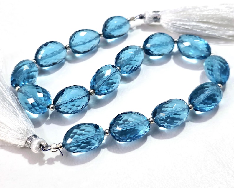 May include: Strand of faceted, oval-shaped blue gemstones. Each gem is connected by a small silver bead. White tassels are attached to each end of the strand. The gems have a transparent, glassy appearance.