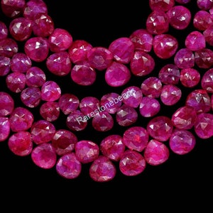 Könnte beinhalten: Eine Strähne aus facettierten Edelsteinperlen in Rubinrot. Die Perlen haben eine tiefe, satte rote Farbe und eine facettierte, fast sternenförmige Optik. Die Perlen sind auf einem durchsichtigen Faden aufgefädelt und bereit für die Schmuckherstellung.