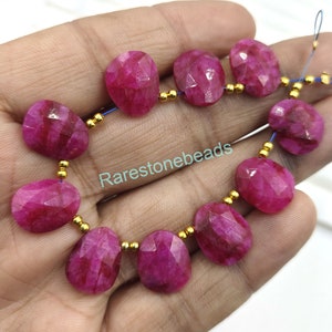 May include: A strand of pink ruby gemstone beads with gold spacers. The beads are oval shaped and have a faceted finish. The beads are strung on a blue thread.