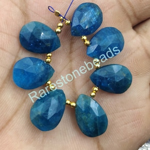 以下が含まれることがあります： 青い宝石のビーズと金のスペーサーのストランド。ビーズは涙滴型で、ファセット加工が施されています。ストランドは青い糸に糸通しされています。Rarestonebeadsが画像に印刷されています。
