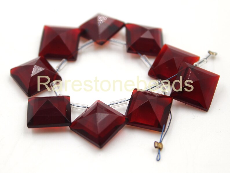 May include: A strand of ten dark red square-shaped gemstone beads with faceted edges. The beads are strung on clear elastic thread.