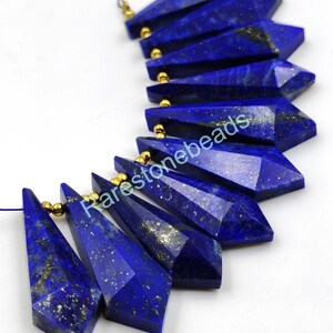 May include: A strand of 10 faceted lapis lazuli gemstone beads with gold-toned spacers. The beads are shaped like long, narrow teardrops with a pointed tip. The lapis lazuli is a deep blue color with flecks of gold pyrite.