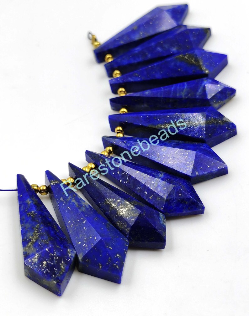May include: A strand of 10 faceted lapis lazuli gemstone beads with gold-toned spacers. The beads are shaped like long, narrow teardrops with a pointed tip. The lapis lazuli is a deep blue color with flecks of gold pyrite.