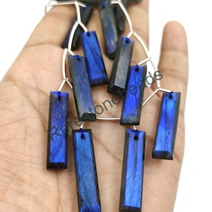 Pode incluir: Um fio de contas de pedra preciosa de labradorita azul escuro, cada conta é um prisma retangular com acabamento facetado. As contas são enfiadas em um fio de prata.