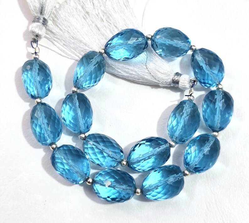 May include: A strand of faceted, oval-shaped, light blue gemstones. The beads are strung together with silver-colored metal beads and a white tassel. The gemstones have a translucent quality, reflecting light.
