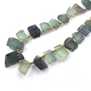 May include: A strand of green and blue fluorite gemstone beads with gold spacers. The beads are irregular in shape and have a natural, rough texture.