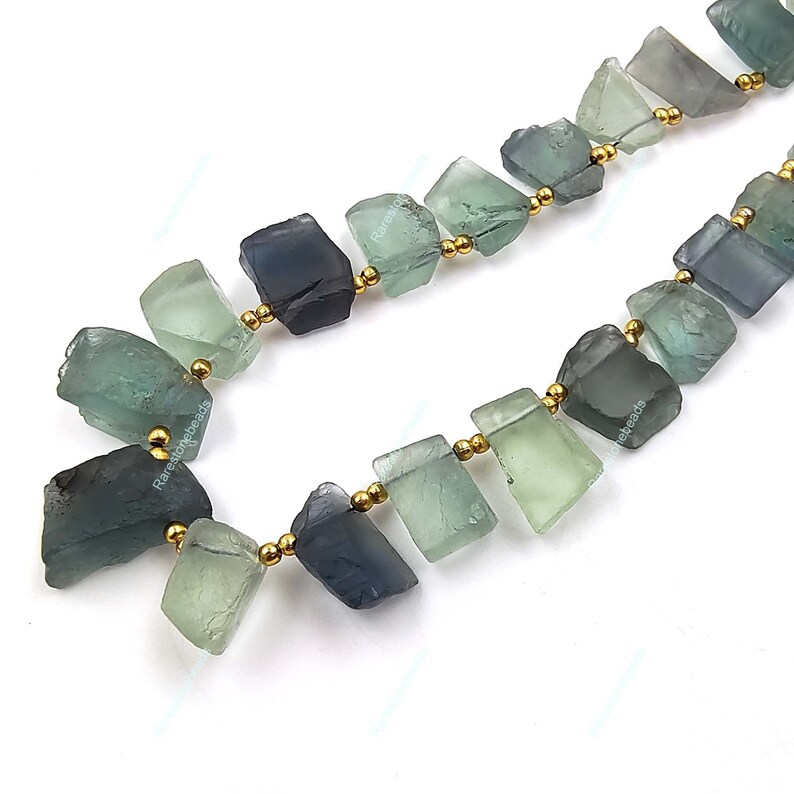 May include: A strand of green and blue fluorite gemstone beads with gold spacers. The beads are irregular in shape and have a natural, rough texture.