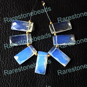 Peut inclure: Un brin de perles de pierre d'opale blanche, chaque perle est rectangulaire et présente un reflet irisé bleu clair. Les perles sont enfilées sur un fil transparent avec des entretoises dorées entre chaque perle.