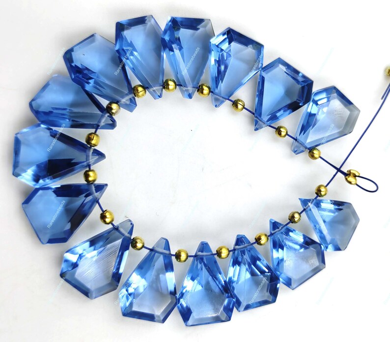 May include: A strand of blue gemstone beads with gold spacers. The beads are faceted and have a teardrop shape. The beads are strung on a clear elastic cord.
