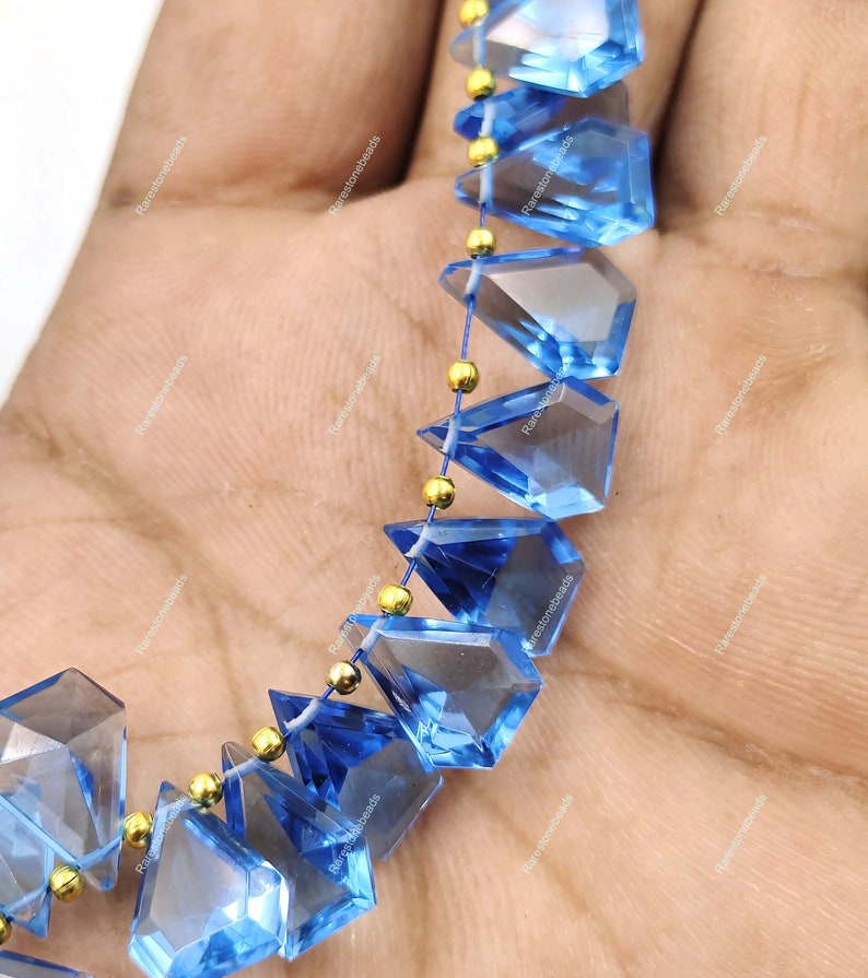 May include: A strand of faceted blue topaz gemstone beads with gold spacers. The beads are faceted in a triangular shape and have a clear, translucent appearance.
