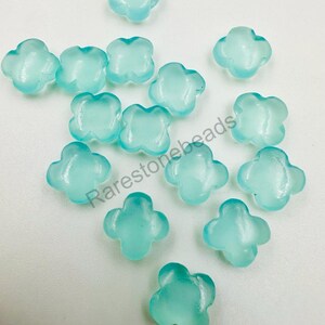 May include: A scattering of light blue, four-petal flower-shaped beads. The beads are spread across a white surface. The text "Rarestonebeads" is visible in the image.