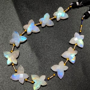 Puede incluir: Una hebra de cuentas de piedra lunar arcoíris iridiscente, cada una tallada en forma de mariposa, enhebradas en un cordón plateado con separadores dorados. Las cuentas están enhebradas en un cordón con un borla negra.