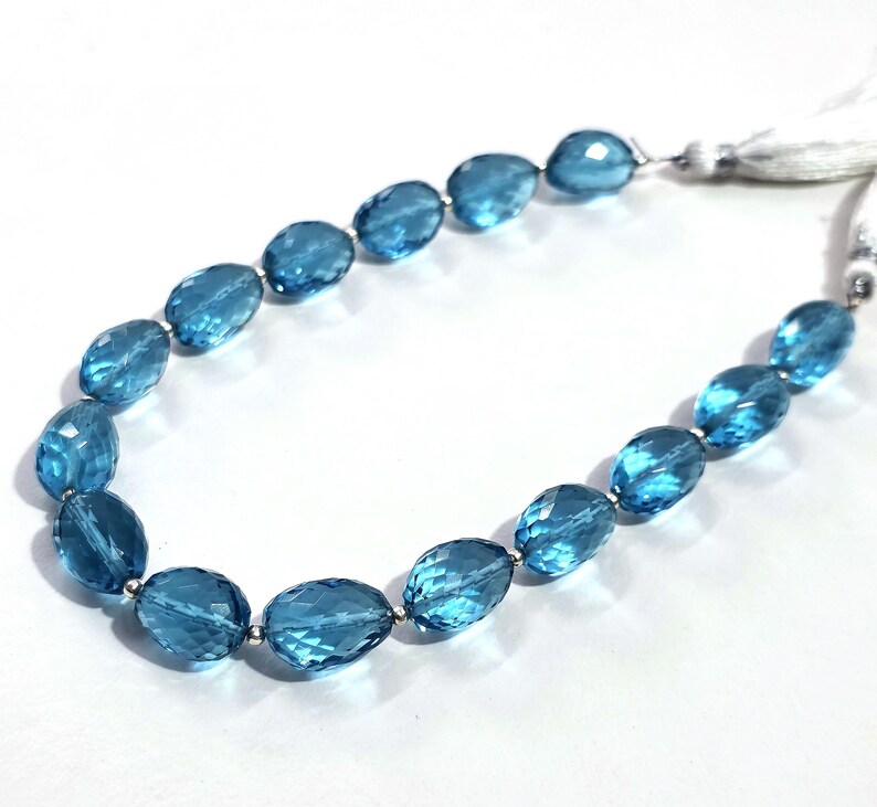May include: A strand of faceted, oval-shaped blue gemstones, strung together with silver-colored beads. The gemstones are a vibrant, transparent blue. The strand ends with a white tassel.