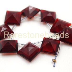 May include: A strand of ten dark red square-shaped gemstone beads with faceted edges. The beads are strung on clear elastic thread.
