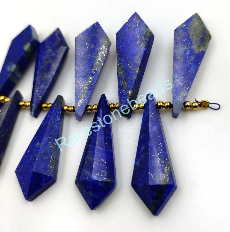 May include: A strand of dark blue lapis lazuli gemstone beads with gold accents. The beads are faceted and have a teardrop shape. The beads are strung on a gold wire.