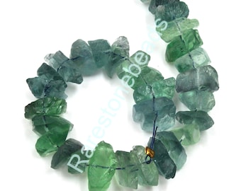 Op de afbeelding: Een streng groene en blauwe fluorite edelsteen kralen. De kralen zijn onregelmatig van vorm en hebben een natuurlijke, ruwe textuur. De kralen zijn geregen op een blauwe draad.