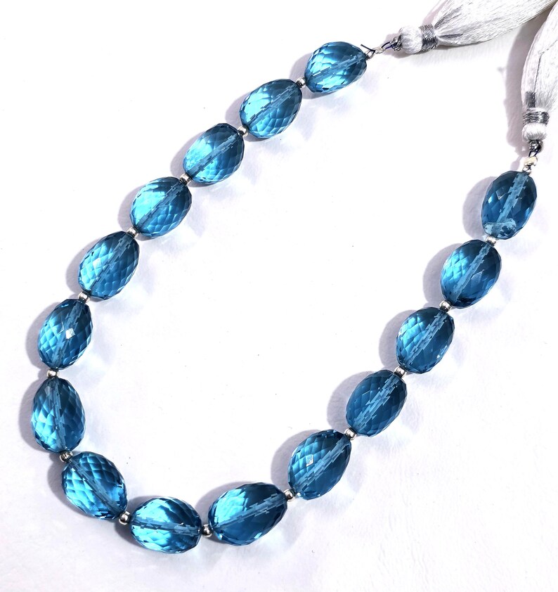 May include: A necklace featuring faceted, oval-shaped blue gemstones strung together with silver-toned metal accents. The necklace has a white tassel closure on each end, creating a delicate and elegant appearance.