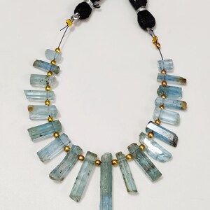 May include: A necklace featuring raw, rectangular aquamarine gemstones in varying shades of blue and green. The stones are strung together with gold-colored beads and black cord, with black tassels at the top.
