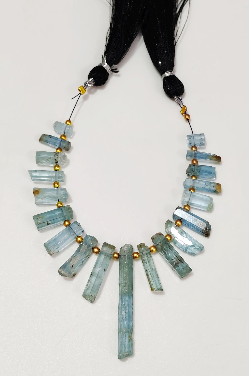 May include: A necklace featuring raw, rectangular aquamarine gemstones in varying shades of blue and green. The stones are strung together with gold-colored beads and black cord, with black tassels at the top.
