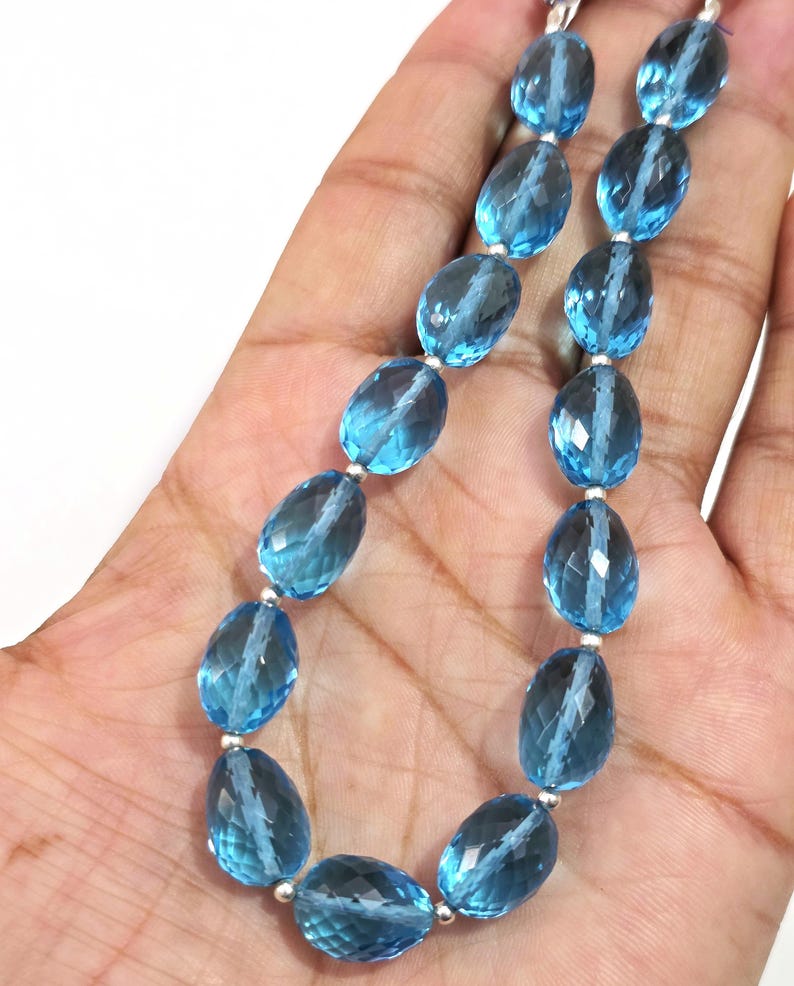May include: A necklace featuring faceted, oval-shaped blue gemstones strung together with silver-colored beads. The gemstones have a translucent quality, and the necklace is displayed against a neutral background.