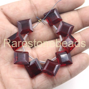 May include: A strand of ten dark red square-shaped glass beads with a faceted finish. The beads are strung on clear thread with silver-toned metal spacers.