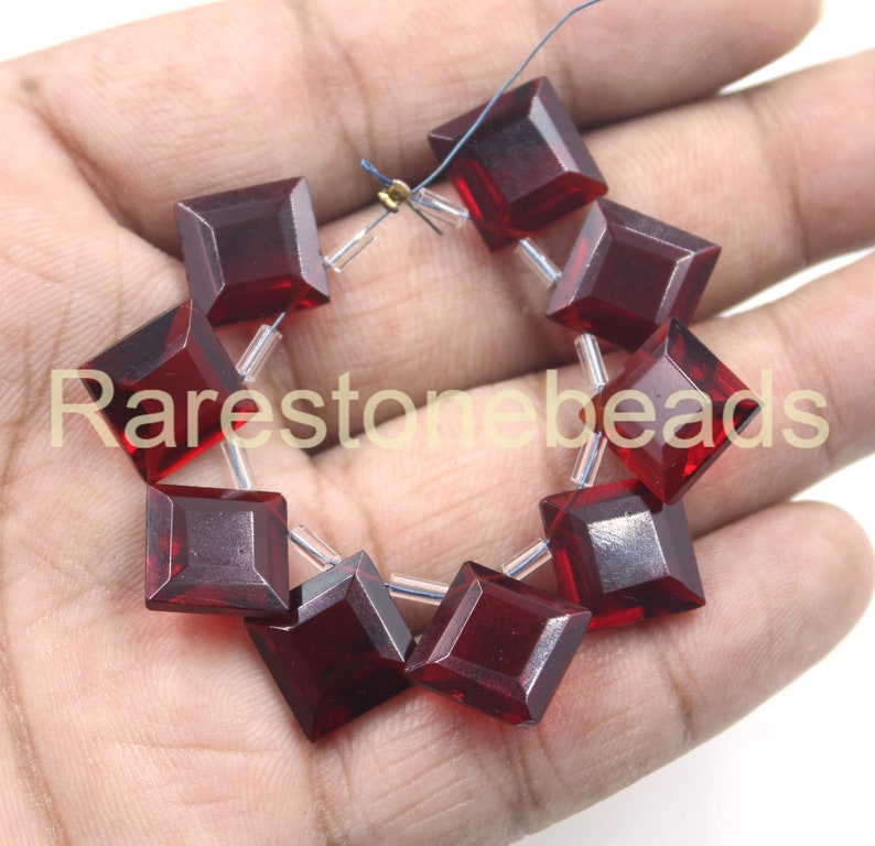 May include: A strand of ten dark red square-shaped glass beads with a faceted finish. The beads are strung on clear thread with silver-toned metal spacers.
