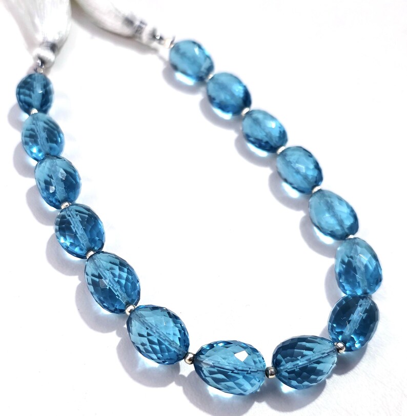May include: A necklace featuring faceted, oval-shaped blue gemstones strung together with silver-colored metal beads. The necklace has a white tassel at the end. The gemstones are a vibrant shade of blue, and the necklace is displayed against a white background.