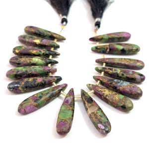 May include: Strand of teardrop-shaped gemstones with a mix of colors including purple, green, and gold. The gemstones are strung together with gold-colored wire and black tassels.
