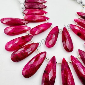 May include: A strand of faceted ruby gemstone beads in a teardrop shape. The beads are a deep red color and are strung on a silver wire.
