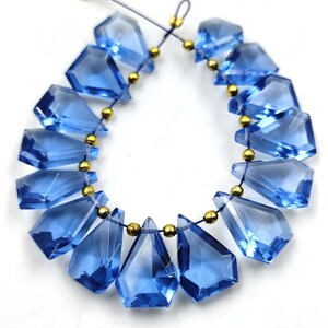 May include: A strand of blue gemstone beads with a faceted, teardrop shape. The beads are strung on a thin cord with gold-colored spacers between each bead.