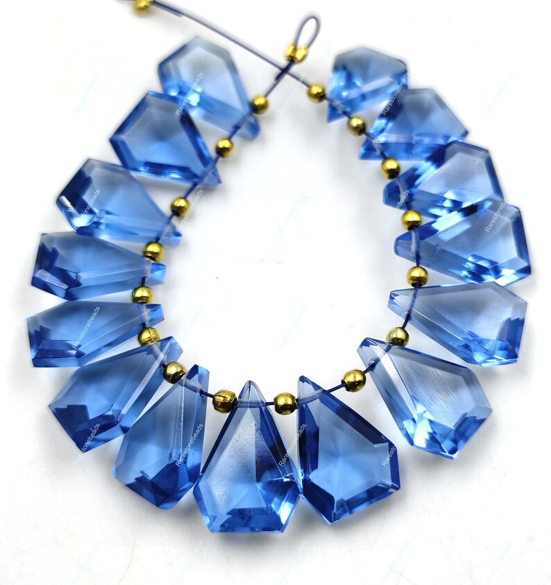 May include: A strand of blue gemstone beads with a faceted, teardrop shape. The beads are strung on a thin cord with gold-colored spacers between each bead.