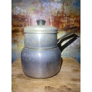 May include: A vintage aluminum coffee pot with a lid and handle. The pot has a cylindrical base and a smaller, lidded upper section. The handle is made of the same metal. The pot is sitting on a wooden surface.