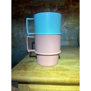 May include: Two stackable mugs with square handles, one in light blue and one in light pink. The mugs feature a smooth, matte-textured finish and a cylindrical shape with a distinct rim where the two pieces connect, creating a vertical stack.