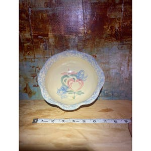 May include: A ceramic bowl with a scalloped edge, featuring a hand-painted design of hearts, flowers, and leaves in pink, green, and blue. The bowl has a light yellow interior and a blue rim. A measuring tape is visible.