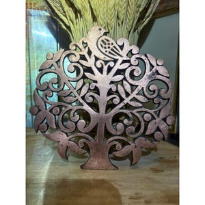 May include: A circular copper-toned metal trivet featuring an intricate openwork design of a tree with swirling branches, leaves, and a bird perched at the top. The metal surface displays a weathered, matte finish with dark patina accents.
