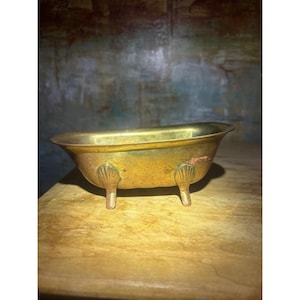 May include: A small, vintage-style brass bathtub dish with a polished metallic finish. The vessel features four decorative feet, each molded into a distinct shell shape, supporting the elongated, curved basin structure of the miniature tub.