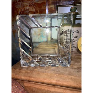 May include: A square clear glass vase featuring a thick, heavy base with a textured, diagonal ribbed pattern along the sides and a smooth, open center. The glass is transparent and reflects light, showcasing a geometric design.