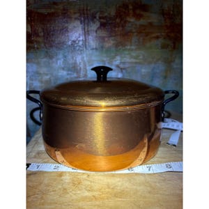 May include: A round copper pot with a matching lid and two black side handles. A white measuring tape is positioned along the base, showing a diameter of approximately 8 inches. The pot has a polished metallic finish and a black knob on the lid.