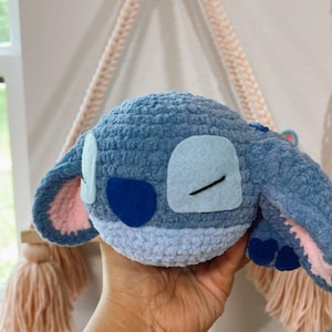 Pode incluir: Um brinquedo de pelúcia azul feito à mão em crochê, semelhante a um personagem de desenho animado. O brinquedo tem orelhas grandes com detalhes internos rosa, manchas azuis para os olhos e um nariz azul escuro. A pelúcia é segurada em uma mão.