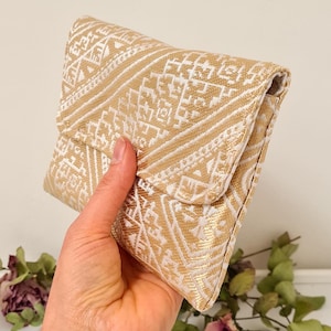 Könnte beinhalten: Eine gold-weiß gemusterte Clutch mit Klappenverschluss. Die Clutch hat ein geometrisches Design und wird in einer Hand gehalten. Der Hintergrund ist eine neutrale Farbe mit getrockneten Blumen.