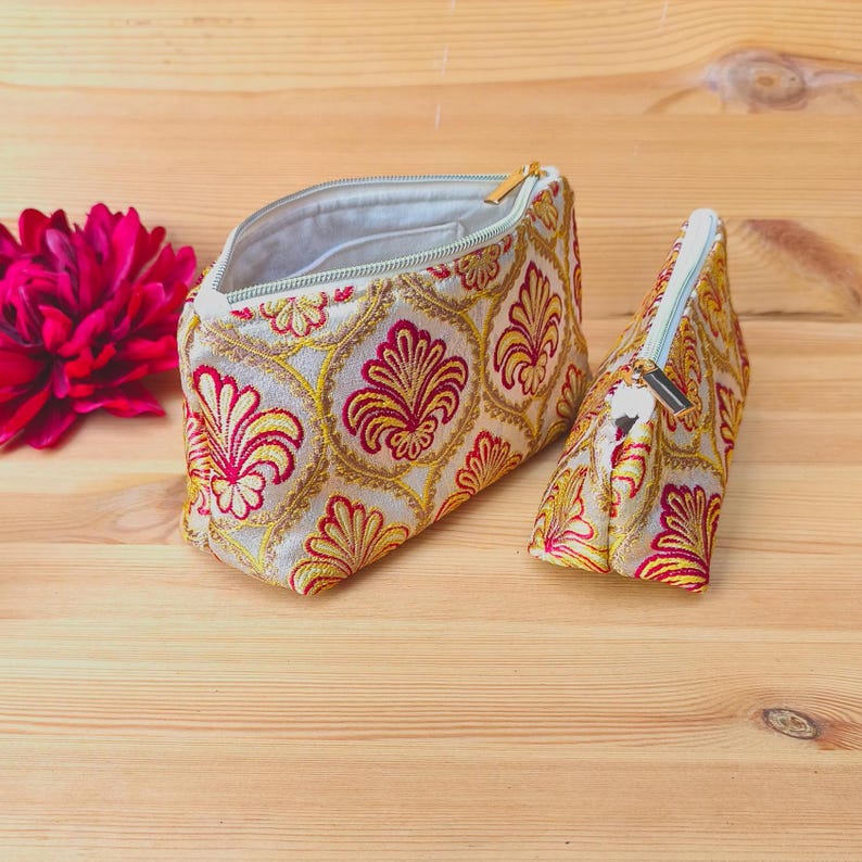 May include: Two beige cosmetic bags with a gold, red, and yellow floral pattern. One bag is open, revealing a light interior. The bags have gold zippers. A bright pink flower is in the background.