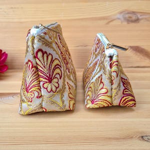 May include: Two decorative cosmetic bags with a gold, red, and yellow floral pattern. The bags have a triangular shape and a zipper closure. The bags are made of a textured fabric and are sitting on a wooden surface.