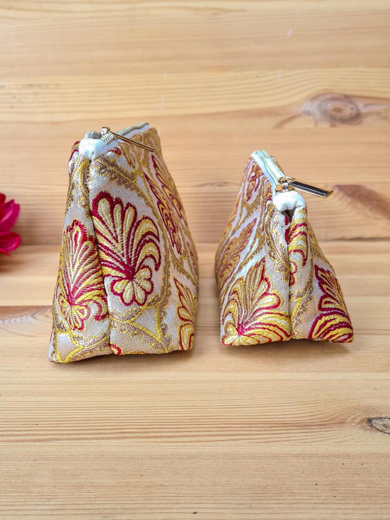 May include: Two decorative cosmetic bags with a gold, red, and yellow floral pattern. The bags have a triangular shape and a zipper closure. The bags are made of a textured fabric and are sitting on a wooden surface.
