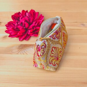 May include: An open, beige cosmetic bag with a gold zipper and intricate red and yellow floral embroidery. A vibrant pink flower is next to the bag on a wooden surface. The bag appears to contain a small orange bottle.
