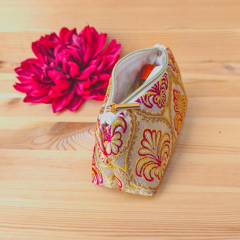 May include: An open, beige cosmetic bag with a gold zipper and intricate red and yellow floral embroidery. A vibrant pink flower is next to the bag on a wooden surface. The bag appears to contain a small orange bottle.