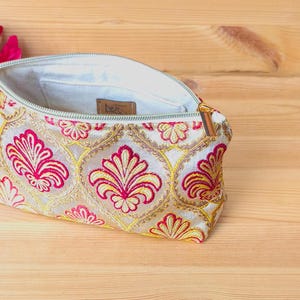 May include: Cream-colored embroidered clutch with a floral pattern in red and gold. The open zipper reveals a light-colored interior. A red flower is visible in the background. The clutch is on a wooden surface.