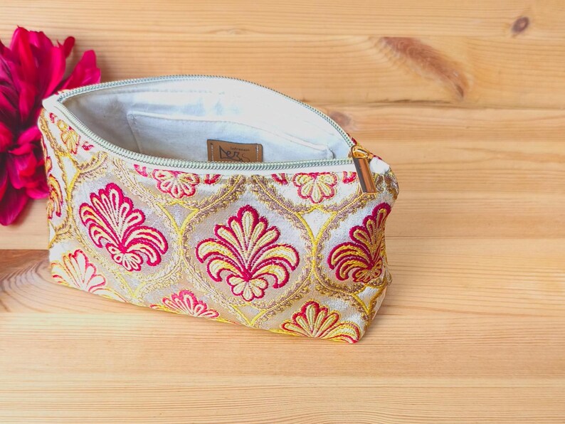 May include: Cream-colored embroidered clutch with a floral pattern in red and gold. The open zipper reveals a light-colored interior. A red flower is visible in the background. The clutch is on a wooden surface.