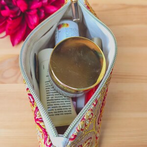 May include: A cosmetic bag with a gold zipper, filled with various beauty products. The bag is decorated with a red and gold floral pattern. A large, dark pink flower is in the background.