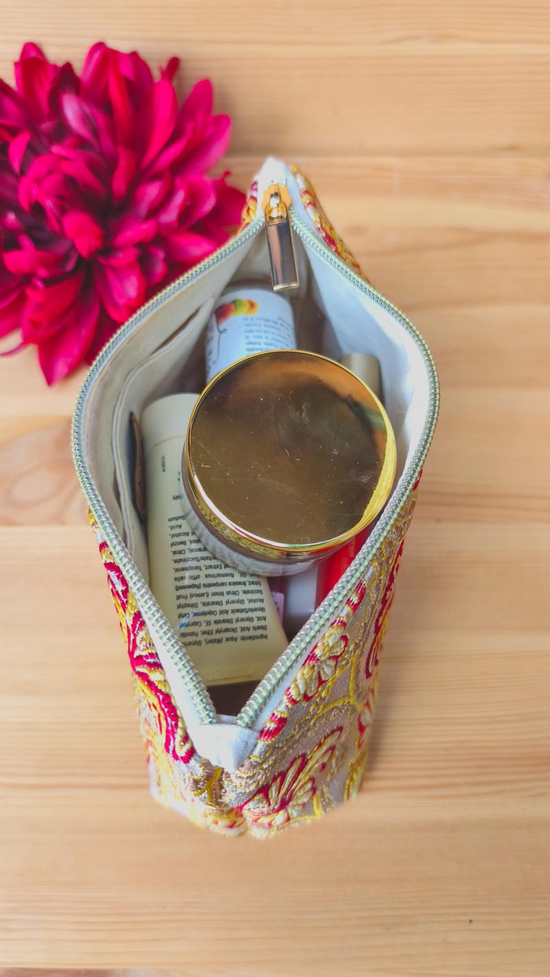 May include: A cosmetic bag with a gold zipper, filled with various beauty products. The bag is decorated with a red and gold floral pattern. A large, dark pink flower is in the background.