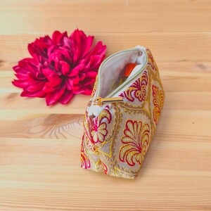 May include: A beige cosmetic bag with a gold zipper, embroidered with red and yellow floral patterns. A large, vibrant pink flower sits beside the open bag. The bag contains a small orange bottle.